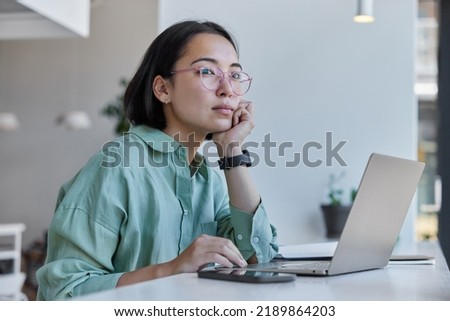 Thoughtful Asian woman works freelancer uses laptop computer smartphone has distance job poses against cozy interior indoors wears spectacles and shirt. Female digital nomad enjoys modern lifestyle Royalty-Free Stock Photo #2189864203