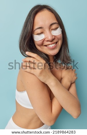 Positive good looking young woman with eastern appearance crosses arms over body touches shoulders gently keeps eyes closed applies beauty patches under eyes dressed in cropped top poses indoor