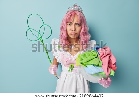 Displeased pink haired Asian housewife dressed like chambermaid holds basin of colorful laundry and rug beater feels tired of domestic duties does regular chores poses indoor against blue background