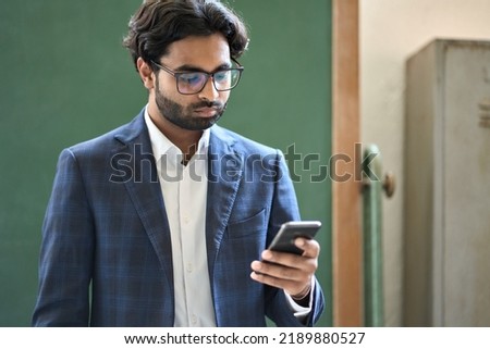 Busy young adult indian business man manager wearing suit holding cell phone working standing in office at work using mobile applications business technology on smartphone checking data, reading