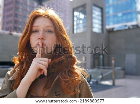 Hipster teen stylish cool redhead fashion girl model showing shh sign asking to keep secret, be hush silent or privacy silence standing in big city urban location. Headshot portrait Royalty-Free Stock Photo #2189880577