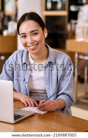 Young smiling Asian woman student using laptop wearing earbud, taking notes online elearning webinars, classes, trainings, remote studying virtual lessons in internet looking at camera. Vertical Royalty-Free Stock Photo #2189887005
