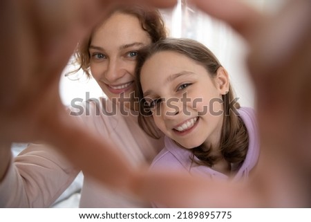 Close up portrait of smiling Caucasian mother and teenage daughter make heart love hand gesture for picture together. Happy mom and teen girl child have fun take selfie at home. Motherhood concept.