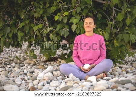 Pleasant woman meditation at summer park sitting on stones with closed eyes mindfulness spiritual balance. Happy yogi female in lotus position enjoy healthy lifestyle near green tree foliage garden