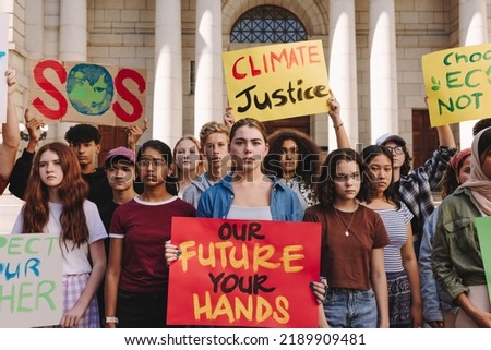Multicultural teenagers holding a climate change protest in the city. Group of youth activists holding banners and placards while marching for climate justice and environmental sustainability.