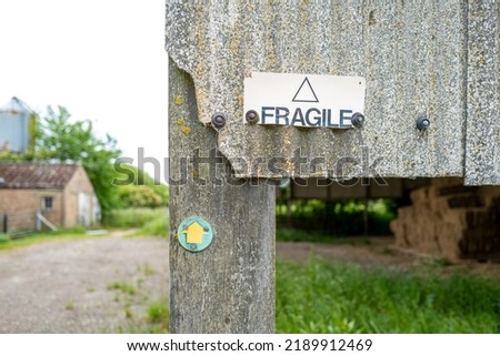 Shallow focus of an old asbestos built farmyard barn showing a Fragile sign. The directional sign is for a public footpath across the farm itself.