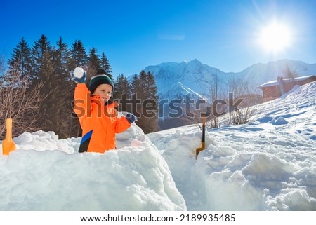 Happy little boy throw snowball standing in the snow fortress with Alps mountains on background during winter vacations