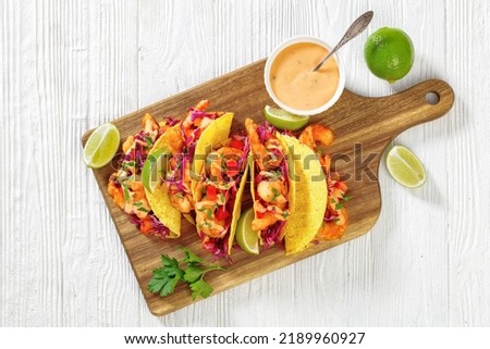 Bang Bang Shrimp Tacos with purple cabbage, tomatoes, parsley, lime drizzled with mayonnaise hot chili sauce on cutting board on white wooden table, horizontal view from above, flat lay