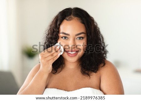 Skincare concept. Happy black overweight lady cleansing face with cotton pad, making self-care beauty routine, standing in light bathroom interior