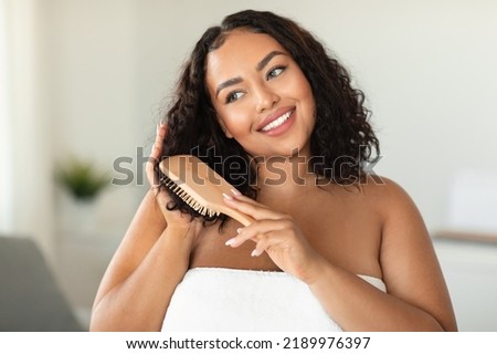 Domestic haircare. Happy african american bodypositive lady brushing hair with brush, standing in bathroom interior, wrapped in towel. Hair beauty care, combing and detangling concept Royalty-Free Stock Photo #2189976397