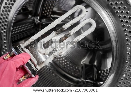 A man holds in his hand a burnt-out heating element of a washing machine against the background of a washing machine drum