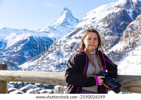 Smiling female traveler enjoying hiking in Swiss Alps in wintertime, standing near wooden fencing in mountains with camera in hands, ready to take pictures