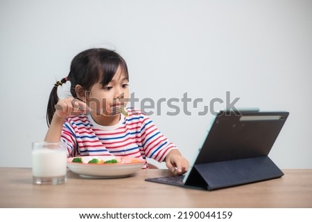 Adorable Asia child girl having lunch while watching a movie from the tablet. A little Asian child eating dinner and eyes are looking cartoon from Tablet. National Eating Disorders Awareness Week.