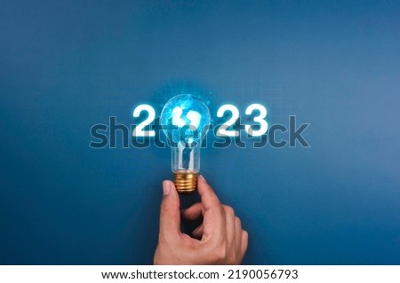 Shining 2023 calendar year numbers, neon style with creative trend light bulb holding by businessman's hand on digital network and blue background. Happy 2022 New Year greeting card with light bulb. Royalty-Free Stock Photo #2190056793