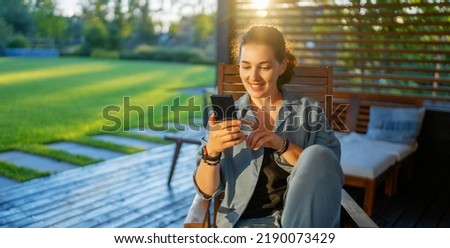 Happy young woman is using a phone sitting on the patio in summer. 