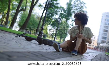 Biracial woman falling down from e-scooter, sitting on ground and rubbing knee Royalty-Free Stock Photo #2190087693