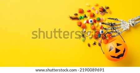 Halloween decorations concept. Top view photo of skeleton hand holding pumpkin basket for trick-or-treat with candies eyes insects centipede and spiders on isolated yellow background