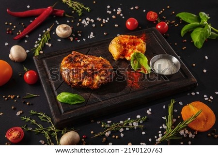 Grilled meat steak in sauce with fried potatoes, basil, sauce and parsley on a wooden board with mushrooms, peas, red chili peppers, salt, tomatoes and basil on a black background