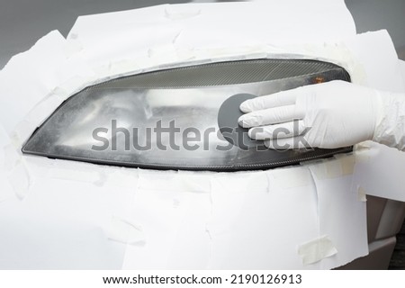 Repairman hand in white rubber protective gloves using sandpaper and sanding car headlight. Renovation process. Restore automobile lamp. Closeup. Front view. Royalty-Free Stock Photo #2190126913