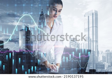 Attractive young european businesswoman using laptop while leaning on desk with creative growing financial forex chart graph on blurry bright city background. Market, stock and trading concept. 
