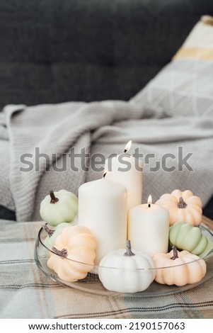 Autumn, fall cozy mood decor composition for hygge home. Decorative pumpkins and white burning candles on the glass tray with gray plaid and sofa background. Vertical card. Selective focus.