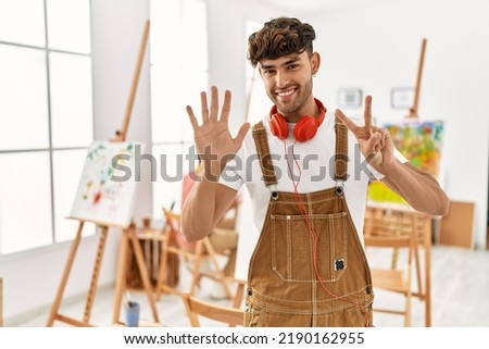 Young hispanic man at art studio showing and pointing up with fingers number seven while smiling confident and happy. 