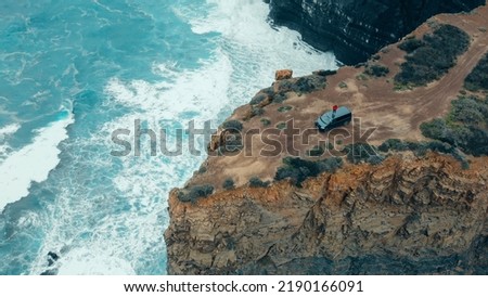 Adventurous woman in red sweater stand on roof of her camper van, make video for social media on smartphone. Aerial shot of travel blogger in epic cinematic location on ocean cliffs.Wanderlust Royalty-Free Stock Photo #2190166091