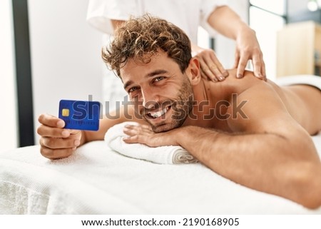 Young hispanic man having back massage holding credit card at beauty center