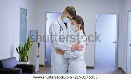 Great looking couple of doctors with a protective mask in the middle of the hospital corridor standing in front of the camera and hugging each other concept of Coronavirus 2019