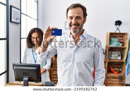 Hispanic man holding credit card at retail shop looking positive and happy standing and smiling with a confident smile showing teeth 