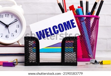 The office desk has diaries, an alarm clock, stationery, and a white card with the text WELCOME. Business concept.