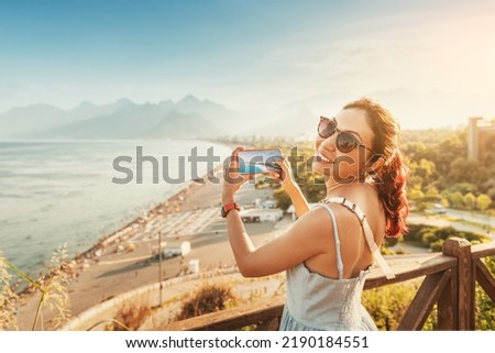 Travel blogger girl takes high-quality and vivid photos on the camera of his new expensive smartphone of the famous Konyaalti beach from a scenic viewpoint in Antalya, Turkey