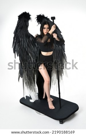 full length portrait of beautiful asian model with dark hair, wearing black gothic skirt costume, angel feather wings with horned headdress. Standing pose  isolated on studio background.