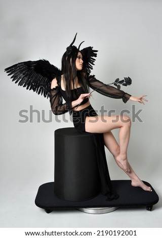 full length portrait of beautiful asian model with dark hair, wearing black gothic skirt costume, angel feather wings with horned headdress. Sitting pose  isolated on studio background.