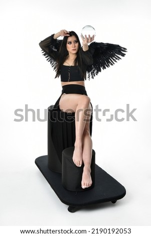 full length portrait of beautiful asian model with dark hair, wearing black gothic skirt costume, angel feather wings with horned headdress. Sitting pose  isolated on studio background.