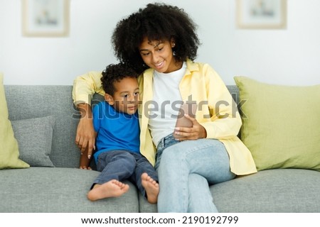 Little ethnic boy watching cartoons on phone smartphone with mother while sitting on couch at home together