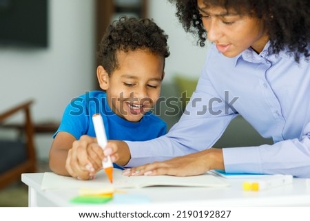 Female infant school teacher working one on one with a young schoolboy, sitting at a table writing in a classroom, front view