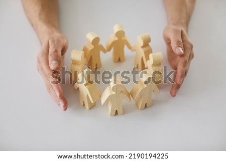 Human hands protecting and guarding small wooden toy figures placed on white desk as metaphor for creating safe, supportive community of people. Close up, closeup shot. Care, support, safety