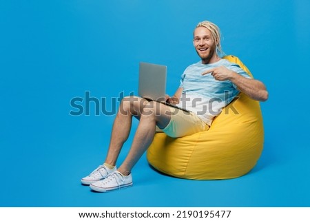 Full body smiling happy young blond man with dreadlocks 20s he wear white t-shirt sit in bag chair hold use work point index finger on laptop pc computer isolated on plain pastel light blue background