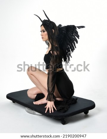full length portrait of beautiful asian model with dark hair, wearing black gothic skirt costume, angel feather wings with horned headdress. kneeling pose  isolated on studio background.