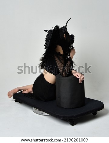 full length portrait of beautiful asian model with dark hair, wearing black gothic skirt costume, angel feather wings with horned headdress. kneeling pose  isolated on studio background.