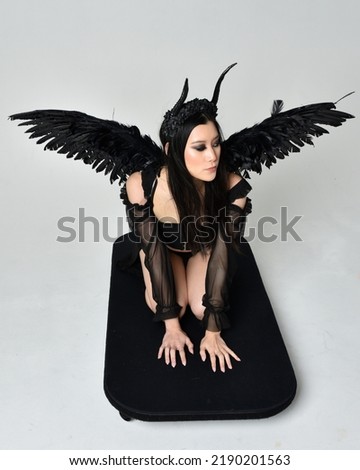 full length portrait of beautiful asian model with dark hair, wearing black gothic skirt costume, angel feather wings with horned headdress. Kneeling pose  isolated on studio background.