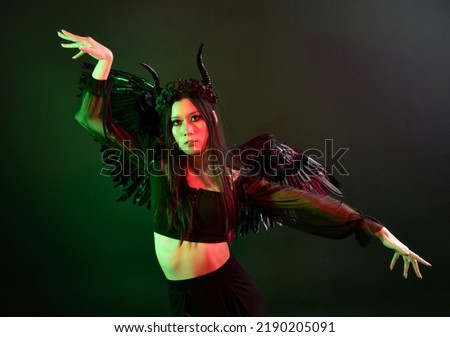 portrait of beautiful asian model with dark hair, wearing black gothic skirt costume, angel feather wings with horned headdress. Posing with gestural hands  on dark silhouette  studio background.