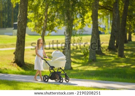 Smiling young adult mother in dress pushing white baby stroller and walking on sidewalk at town park in warm sunny summer day. Spending time with newborn and breathing fresh air. Enjoying stroll. Royalty-Free Stock Photo #2190208251