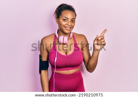 Young african american girl wearing gym clothes and using headphones with a big smile on face, pointing with hand finger to the side looking at the camera. 