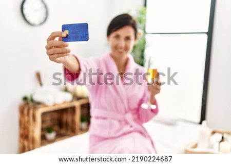 Middle age hispanic woman toasting with champagne holding credit card at beauty center