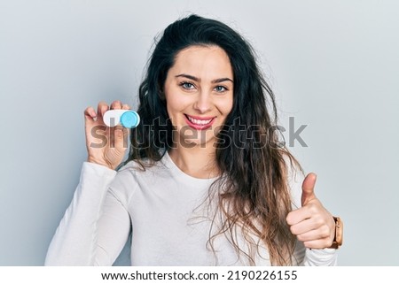 Young hispanic woman holding contact lenses smiling happy and positive, thumb up doing excellent and approval sign 