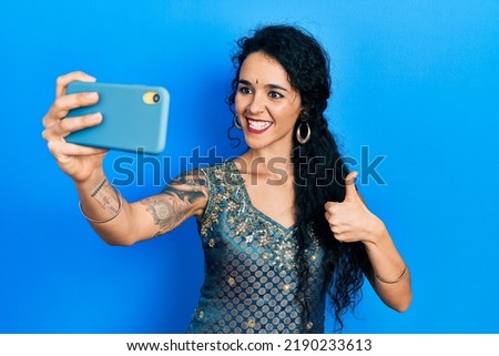 Young woman wearing bindi and traditional kurta dress taking a selfie photo with smartphone smiling happy and positive, thumb up doing excellent and approval sign 