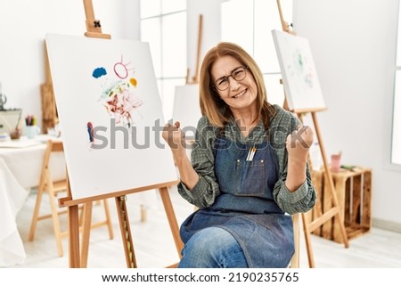 Middle age artist woman at art studio very happy and excited doing winner gesture with arms raised, smiling and screaming for success. celebration concept. 