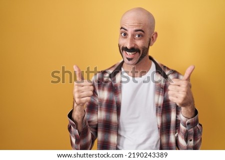 Hispanic man with beard standing over yellow background success sign doing positive gesture with hand, thumbs up smiling and happy. cheerful expression and winner gesture. 